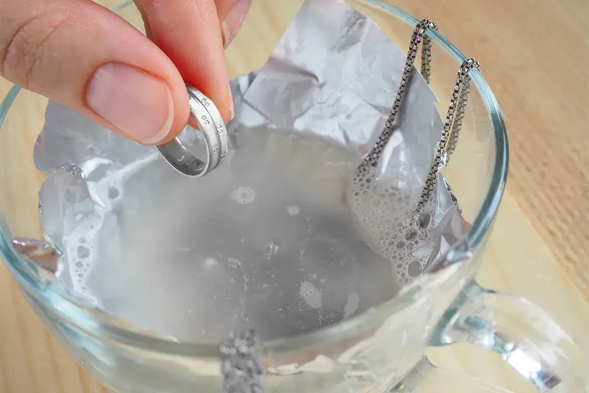 Dłoń wkłada srebrny pierścionek do szklanki z wodą i folią aluminiową, pokazując, jak wyczyścić srebrną biżuterię.